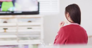 Caucasian woman sitting on couch, eating snacks and watching tv in living room. Spending quality time at home concept.