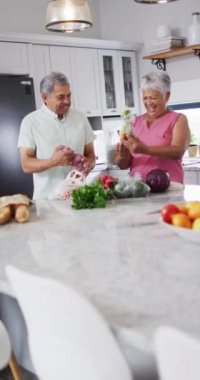 Vertical video of happy senior biracial couple laughing and holding vegetables. Spending quality time at home, retirement and lifestyle concept.