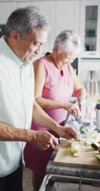 Vertical video of happy senior biracial couple preparing healthy drink. Spending quality time at home, retirement and lifestyle concept.