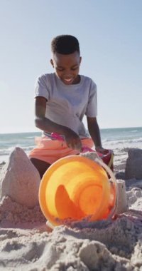 Vertical video of happy african american boy on beach playing with bucket. Holidays, vacations and relax on beach concept.