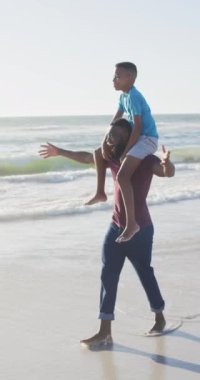 Vertical video of happy african american father and son having fun on beach. Holidays, vacations, summer, family and relationship concept.