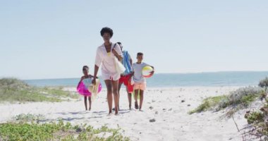 Happy african american family with beach equipment walking on beach in a sunny day. Holidays, vacations, relax and spending quality family time together concept.