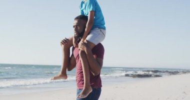 Video of happy african american father holding son on the shoulders at the beach at sunset. Holidays, vacations, relax and spending quality family time together concept.