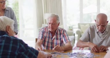 Diverse group of senior male and female friends doing jigsaw puzzle in living room, slow motion. Friendship, relaxation and senior lifestyle.