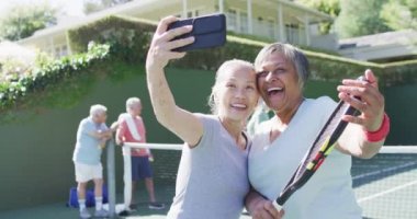Two happy diverse senior women taking selfie at sunny outdoor tennis court with friends, slow motion. Friendship and healthy, active senior lifestyle.