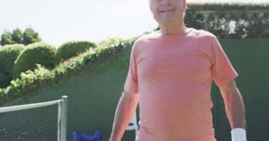 Elinde raket tutan, açık hava tenis kortuna gülen, ağır çekimde mutlu beyaz adam. Arkadaşlık ve sağlıklı, aktif kıdemli yaşam tarzı.