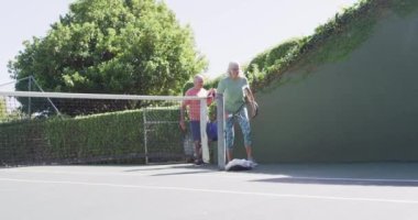 Smiling senior caucasian couple, arriving to play tennis at sunny outdoor tennis court, slow motion. Friendship and healthy, active senior lifestyle.