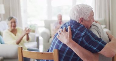 Two happy diverse senior men hugging in living room, with diverse senior friends, slow motion. Friendship, support and senior lifestyle.