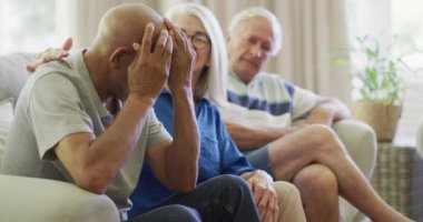 Senior caucasian couple consoling sad diverse senior male friend in living room, slow motion. Friendship, support and senior lifestyle.