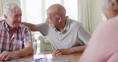 Two diverse senior male friends embracing, doing jigsaw puzzle in living room, slow motion. Friendship, relaxation and senior lifestyle.