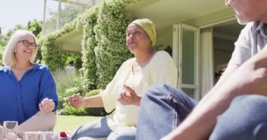 Happy biracial senior woman talking with diverse friends at a picnic in sunny garden, slow motion. Friendship, relaxation and active senior lifestyle.