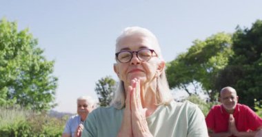Bahçede çeşitli son sınıf öğrencileriyle yoga meditasyonu yapan kıdemli beyaz kadın, ağır çekimde. Sağlıklı, aktif yaşlı yaşam tarzı.
