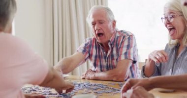 Diverse group of happy senior male and female friends doing jigsaw in living room, slow motion. Friendship, relaxation and senior lifestyle.