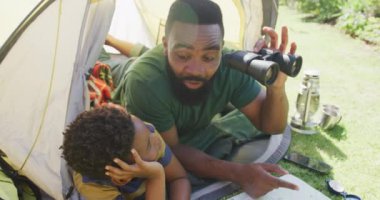 Mutlu Afro-Amerikan baba ve oğlu çadırda oturmuş dürbünle ağır çekimde izliyorlar. Kaliteli zaman geçirme, ev hayatı ve çocukluk kavramı.