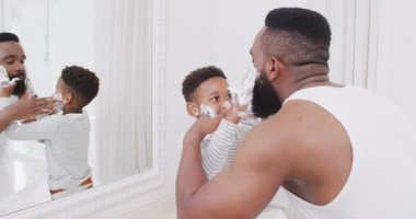 Afro-Amerikan baba ve oğlu banyoda yüzüne tıraş kremi sürüyorlar, yavaş çekimde. Kaliteli zaman geçirme, ev hayatı ve çocukluk kavramı.