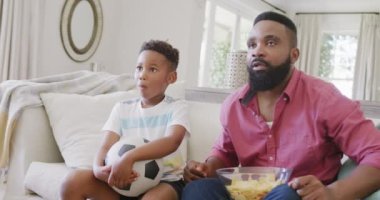 Mutlu Afro-Amerikan baba ve oğlu futbol maçı izliyor ve destekliyor, ağır çekimde. Kaliteli zaman geçirme, ev hayatı ve çocukluk kavramı.