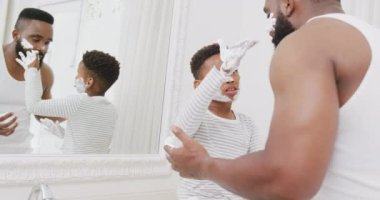 Afro-Amerikan baba ve oğlu banyoda yüzüne tıraş kremi sürüyorlar, yavaş çekimde. Kaliteli zaman geçirme, ev hayatı ve çocukluk kavramı.