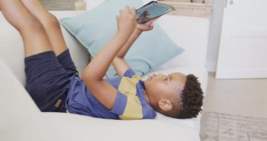 Mutlu Afro-Amerikan çocuk koltukta oturuyor ve tablet kullanıyor, ağır çekimde. Kaliteli zaman geçirme, ev hayatı ve çocukluk kavramı.