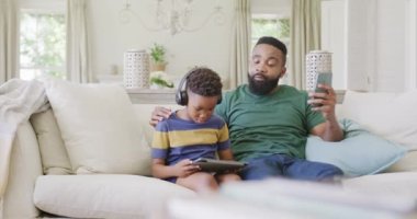 Mutlu Afro-Amerikan baba ve oğlu koltukta oturuyor, akıllı telefon ve tablet kullanıyorlar, ağır çekimde. Kaliteli zaman geçirme, ev hayatı ve çocukluk kavramı.