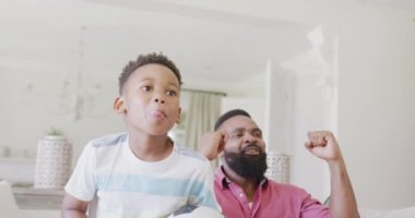 Mutlu Afro-Amerikan baba ve oğlu futbol maçı izliyor ve destekliyor, ağır çekimde. Kaliteli zaman geçirme, ev hayatı ve çocukluk kavramı.