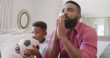 Mutlu Afro-Amerikan baba ve oğlu futbol maçı izliyor ve destekliyor, ağır çekimde. Kaliteli zaman geçirme, ev hayatı ve çocukluk kavramı.