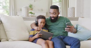 Mutlu Afro-Amerikan baba ve oğlu koltukta oturuyor, akıllı telefon ve tablet kullanıyorlar, ağır çekimde. Kaliteli zaman geçirme, ev hayatı ve çocukluk kavramı.