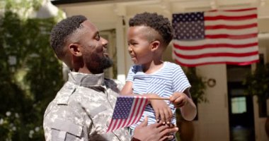 Mutlu Afro-Amerikan erkek askeri yavaş çekimde oğlunu Amerikan bayrağıyla kucaklıyor. Yurtseverlik kavramında kaliteli zaman geçirmek.