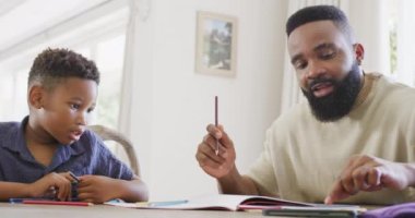 Mutlu Afro-Amerikan baba ve oğlu masada oturup ödevlerini ağır çekimde yapıyorlar. Kaliteli zaman geçirme, ev hayatı ve çocukluk kavramı.