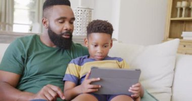 Afro-Amerikan baba ve oğlu kanepede oturup tablet kullanarak ağır çekimde mutlu mesut yaşıyorlar. Kaliteli zaman geçirme, ev hayatı ve çocukluk kavramı.