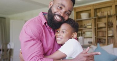 Mutlu Afro-Amerikan baba ve oğlunun portresi koltukta oturuyor ve kucaklaşıyor, ağır çekimde. Kaliteli zaman geçirme, ev hayatı ve çocukluk kavramı.