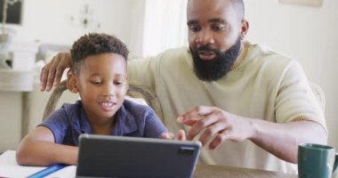 Mutlu Afro-Amerikan baba ve oğlu masada oturup ödevlerini ağır çekimde yapıyorlar. Kaliteli zaman geçirme, ev hayatı ve çocukluk kavramı.