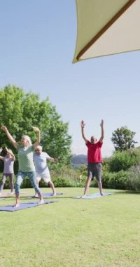 Vertical video of happy diverse senior men and women doing yoga in sunny garden, in slow motion. Healthy, active senior lifestyle.