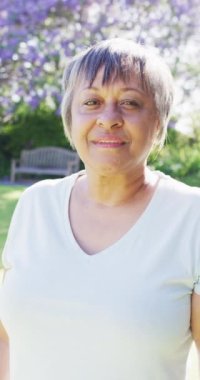 Bahçede yavaşça gülümseyen mutlu, melez bir kadının dikey video portresi. Sağlıklı, aktif yaşlı yaşam tarzı.