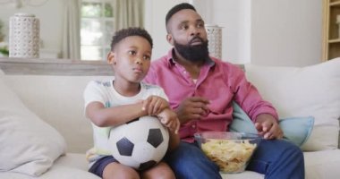 Mutlu Afro-Amerikan baba ve oğlu futbol maçı izleyip cips yiyorlar, ağır çekimde. Kaliteli zaman geçirme, ev hayatı ve çocukluk kavramı.