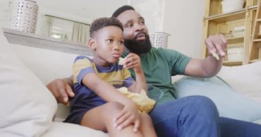 Mutlu Afro-Amerikan baba ve oğlu kanepede oturup televizyon izliyor ve ağır çekimde cips yiyorlar. Kaliteli zaman geçirme, ev hayatı ve çocukluk kavramı.