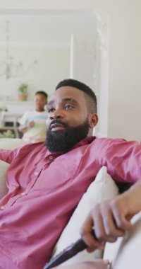 Afrikalı Amerikalı baba ve oğlunun kanepede oturup kucaklaşmalarını gösteren dikey bir video. Kaliteli zaman geçirme, ev hayatı ve çocukluk kavramı.