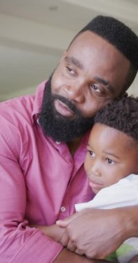 Afrikalı Amerikalı baba ve oğlunun kanepede oturup kucaklaşmalarını gösteren dikey bir video. Kaliteli zaman geçirme, ev hayatı ve çocukluk kavramı.