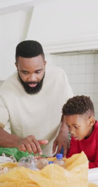 Afrikalı Amerikalı baba ve oğlunun mutfakta yavaş çekimde atık ayıkladıkları dikey video. Kaliteli zaman geçirme, ev hayatı ve çocukluk kavramı.