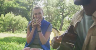 Kamp yapan, çay içen ve parkta gitar çalan mutlu çift, yavaş çekim. Doğa, yürüyüş ve park konsepti.