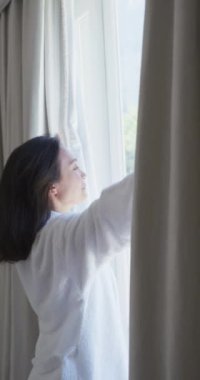 Yatak odasındaki perdeleri yavaş çekimde açan Asyalı kadının dikey videosu. Kaliteli zaman, yaşam tarzı ve ev hayatı kavramı harcamak.