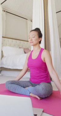 Asyalı kadının yoga yaptığı ve yatak odasında yavaş çekimde meditasyon yaptığı dikey bir video. Kaliteli zaman, yaşam tarzı ve ev hayatı kavramı harcamak.