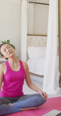 Asyalı kadının yoga yaptığı ve yatak odasında yavaş çekimde meditasyon yaptığı dikey bir video. Kaliteli zaman, yaşam tarzı ve ev hayatı kavramı harcamak.