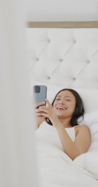 Yatakta uzanan ve yatak odasında ağır çekimde akıllı telefon kullanan mutlu Asyalı kadının dikey videosu. Kaliteli zaman, yaşam tarzı ve ev hayatı kavramı harcamak.