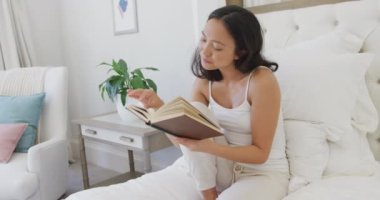 Yatakta oturup kitap okuyan mutlu Asyalı kadın, ağır çekimde. Kaliteli zaman, yaşam tarzı ve ev hayatı kavramı harcamak.