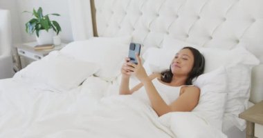 Mutlu Asyalı kadın yatakta uzanıyor, kulaklık takıyor ve akıllı telefon kullanıyor, ağır çekimde. Kaliteli zaman, yaşam tarzı ve ev hayatı kavramı harcamak.