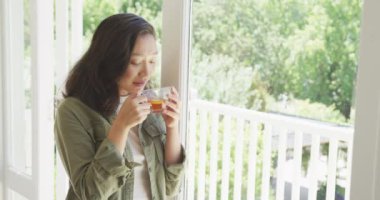 Çay içip pencereden bakan mutlu Asyalı kadın, ağır çekimde. Kaliteli zaman, yaşam tarzı ve ev hayatı kavramı harcamak.