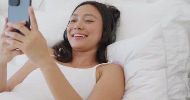 Mutlu Asyalı kadın yatakta uzanıyor, kulaklık takıyor ve akıllı telefon kullanıyor, ağır çekimde. Kaliteli zaman, yaşam tarzı ve ev hayatı kavramı harcamak.