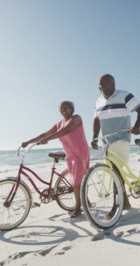 Yaşlı Afro-Amerikan çiftlerin yavaş çekimde sahilde bisikletle yürüdüklerini gösteren dikey bir video. Kaliteli zaman, yaşam tarzı, emeklilik ve tatil konsepti.