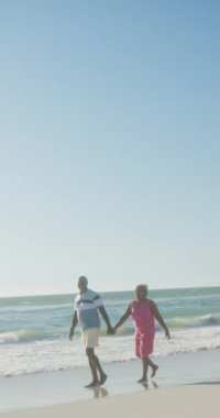 Yaşlı Afro-Amerikan çiftin kumsalda el ele tutuşmalarını gösteren dikey bir video. Kaliteli zaman, yaşam tarzı, emeklilik ve tatil konsepti.