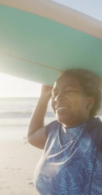 Sahilde sörf tahtasını ağır çekimde tutan mutlu Afrikalı Amerikalı kadının dikey videosu. Kaliteli zaman, yaşam tarzı, emeklilik ve tatil konsepti.